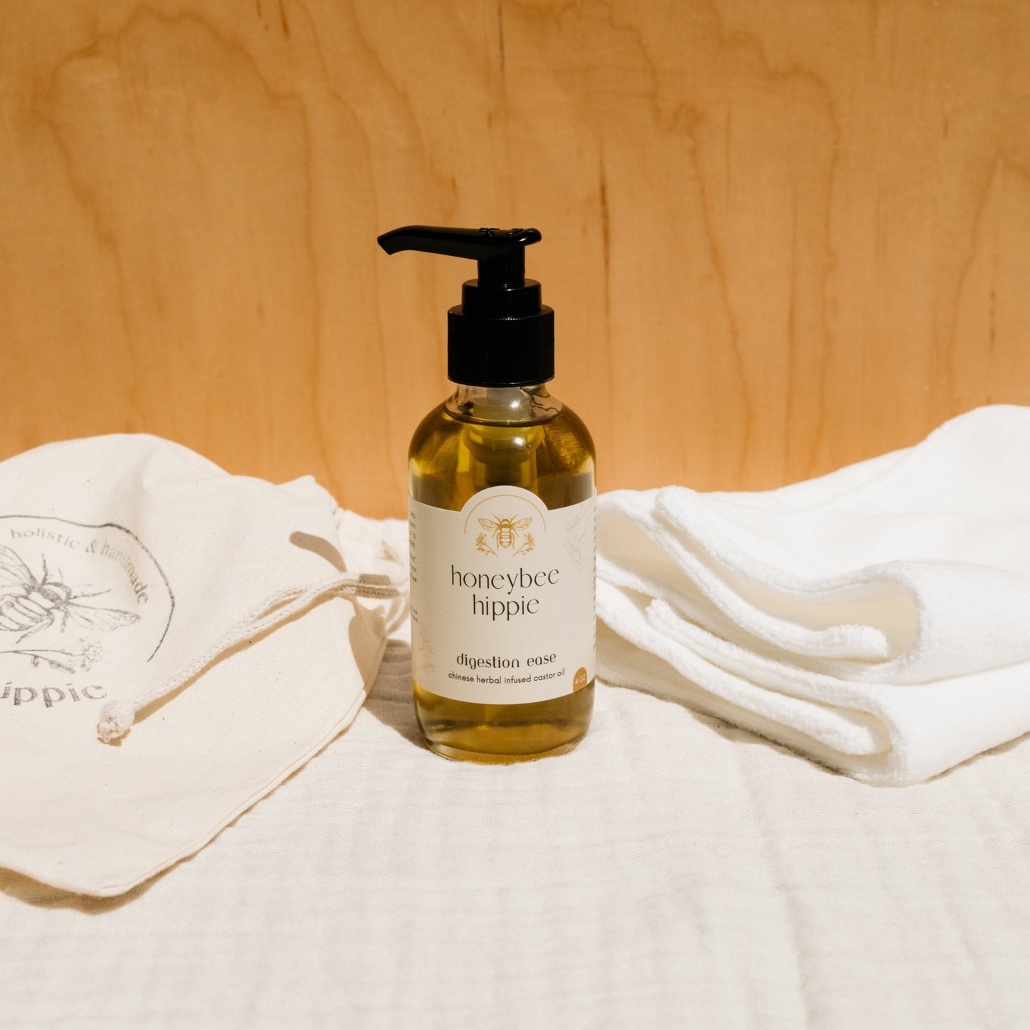 A bottle of Honeybee Hippie's "herbal infused castor oil packs" with a pump top sits on light fabric, surrounded by folded white cloths in a serene setting. Infused with castor oil, it embodies holistic health, enhanced by the warmth of the wooden background.