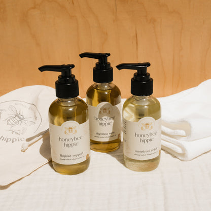 Three Honeybee Hippie herbal-infused castor oil packs with pump dispensers sit against a wooden backdrop, labeled "thyroid support," "digestion ease," and "menstrual relief." Embrace holistic health with these blends. A bee-logo cloth peeks out in the background.