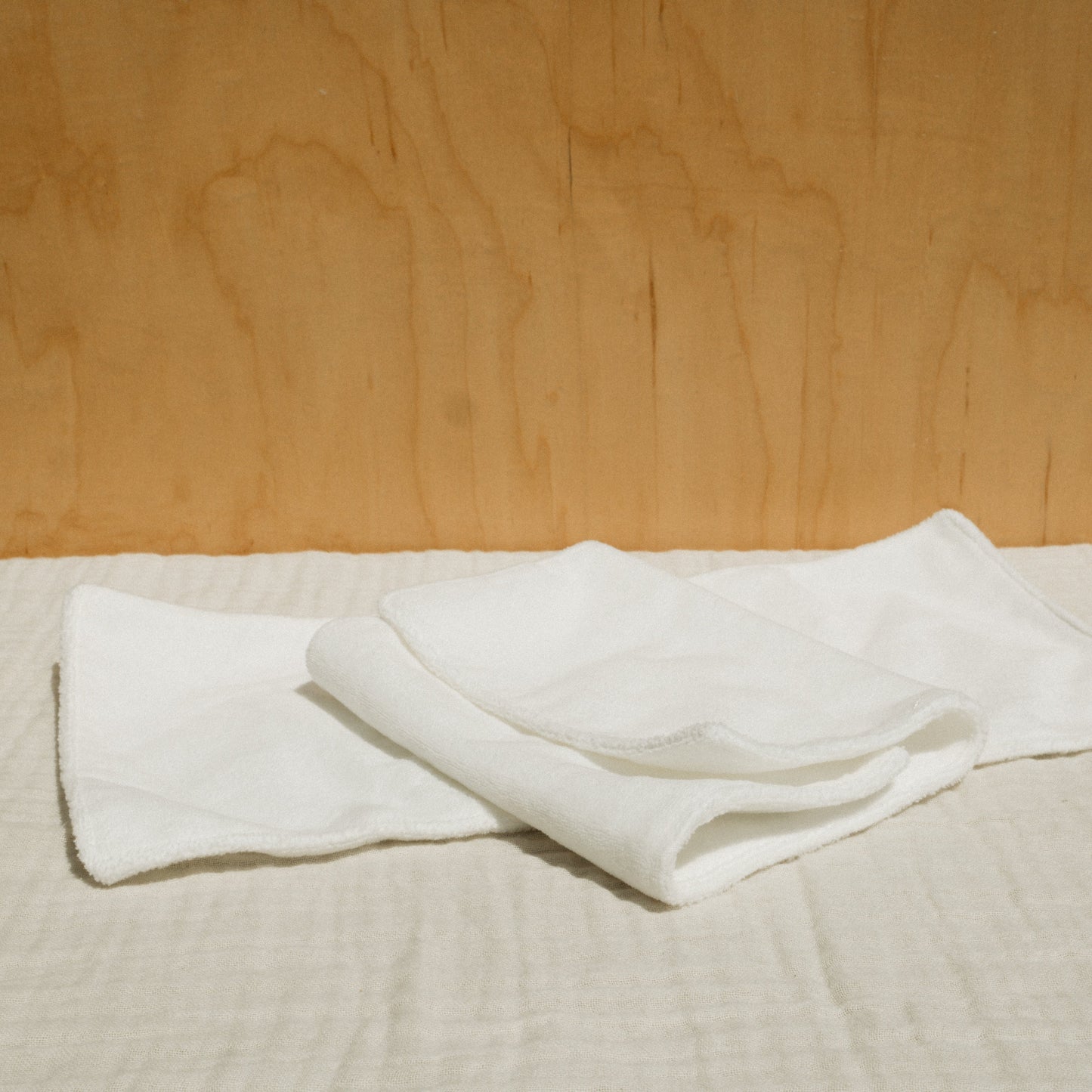 A neatly folded white cloth and a small bottle of Honeybee Hippie's herbal infused castor oil packs rest on checkered beige fabric against a light brown wooden background, creating a softly lit scene that evokes holistic health and tranquility.