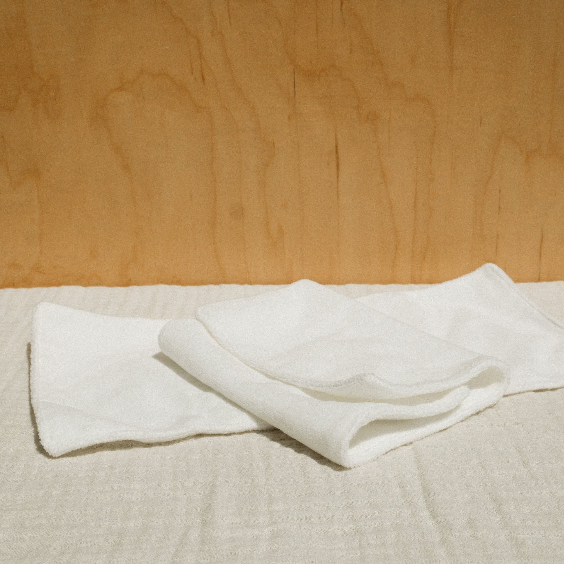 A neatly folded white cloth and a small bottle of Honeybee Hippie's herbal infused castor oil packs rest on checkered beige fabric against a light brown wooden background, creating a softly lit scene that evokes holistic health and tranquility.