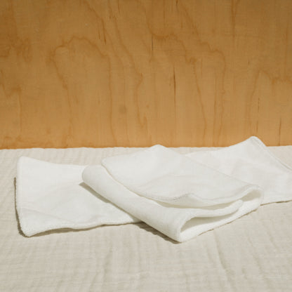 A neatly folded white cloth and a small bottle of Honeybee Hippie's herbal infused castor oil packs rest on checkered beige fabric against a light brown wooden background, creating a softly lit scene that evokes holistic health and tranquility.