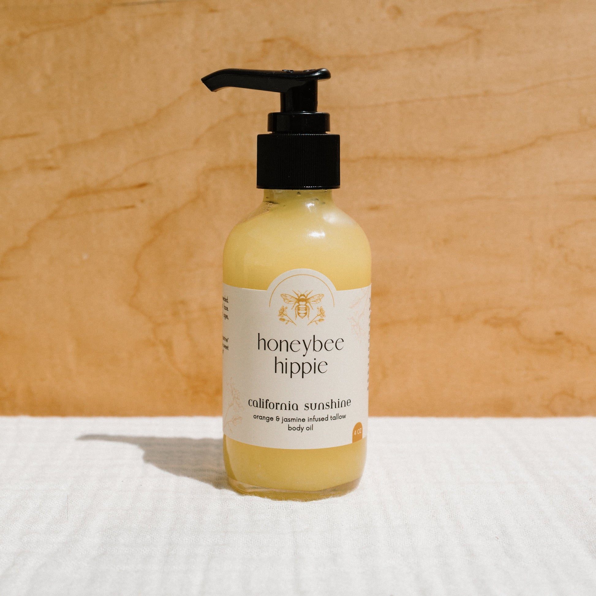 A clear bottle with a black pump holds the yellow "california sunshine - tallow oil" by Honeybee Hippie, featuring organic orange peel and jasmine notes. Non-comedogenic, it sits on a white surface against a light wooden background.
