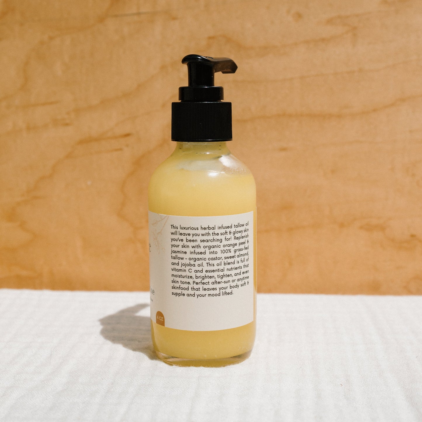 A bottle with a black pump, labeled "California Sunshine - Tallow Oil" by Honeybee Hippie, sits on a textured white surface. Its non-comedogenic oil boasts organic orange peel essence, with the yellow liquid contrasting beautifully against the beige wood-texture background.