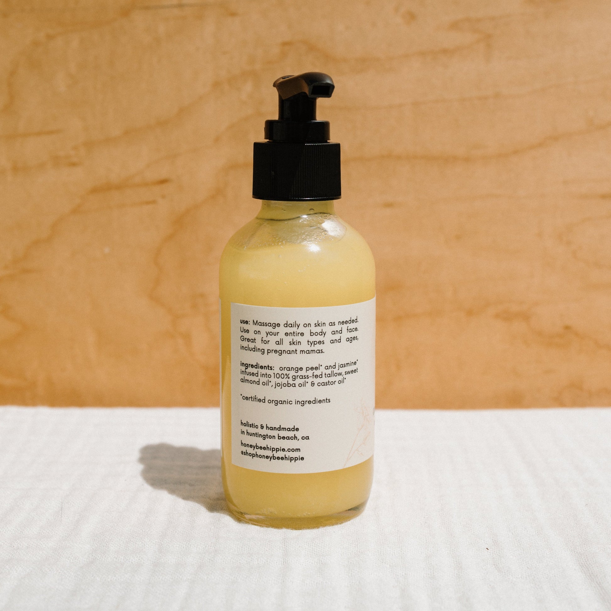 A glass pump bottle of Honeybee Hippie's "california sunshine - tallow oil," containing yellow lotion with organic orange peel, stands on a textured surface against a neutral background. The label lists uses and ingredients, featuring a non-comedogenic formula that nourishes without clogging pores.