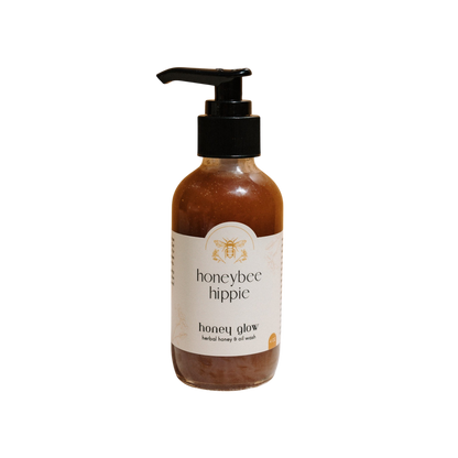 A bottle of Honeybee Hippie's "Honey Glow" herbal honey & oil wash with a pump top features a bee illustration on a white label, highlighting raw honey and organic oils. The amber gel contrasts beautifully against the black backdrop.