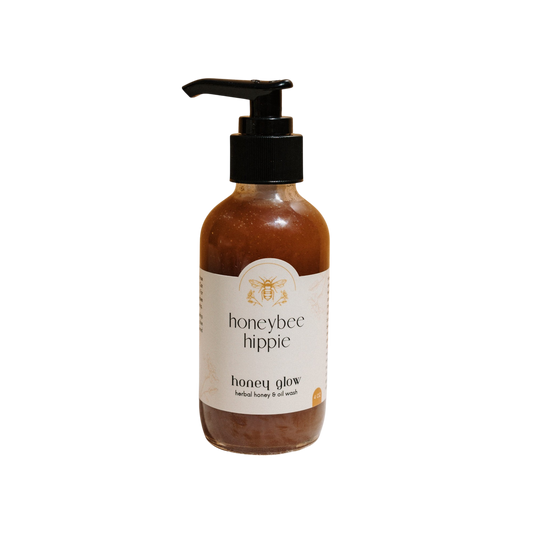 A bottle of Honeybee Hippie's "Honey Glow" herbal honey & oil wash with a pump top features a bee illustration on a white label, highlighting raw honey and organic oils. The amber gel contrasts beautifully against the black backdrop.