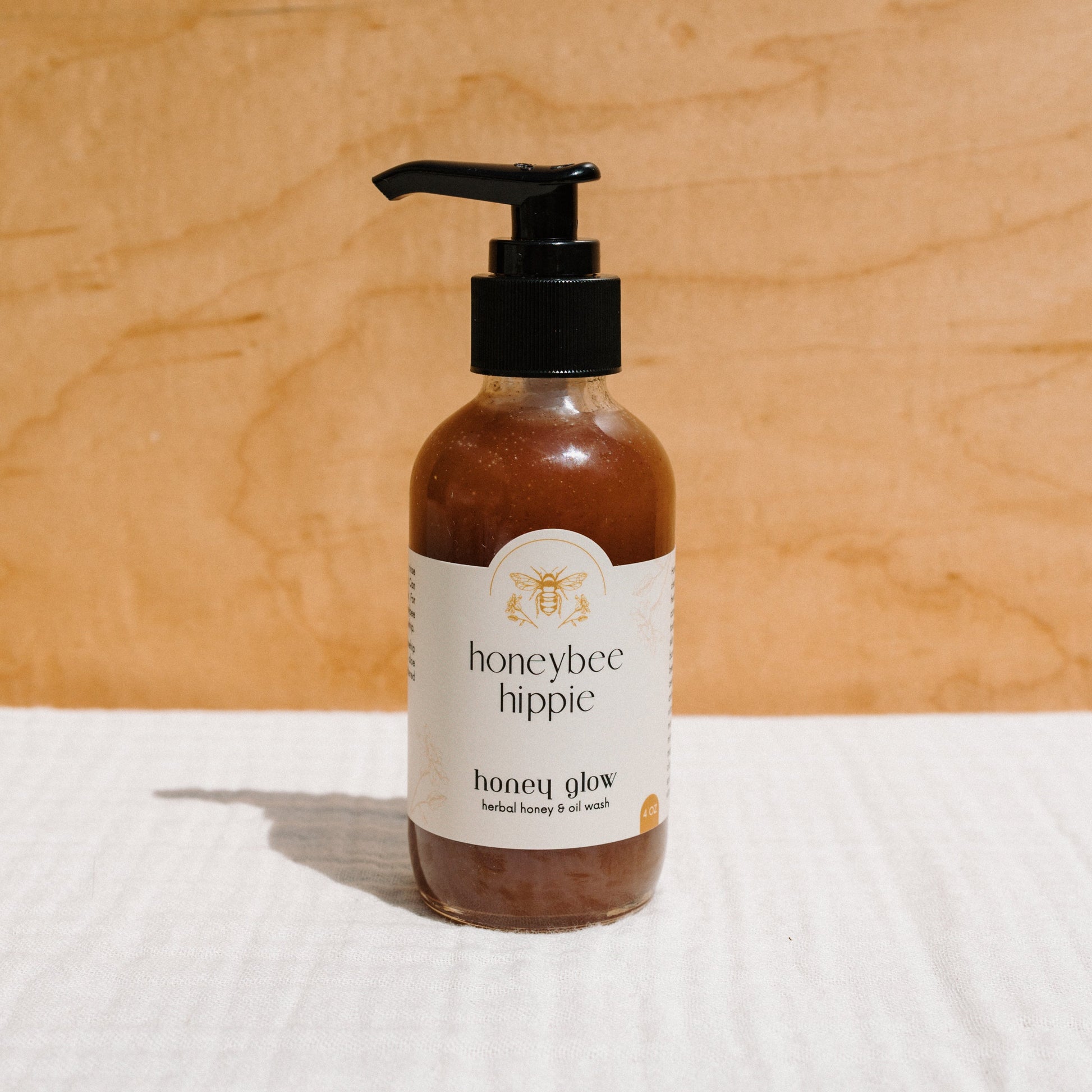 A brown bottle with a black pump labeled "Honeybee Hippie" and "honey glow herbal honey & oil wash" stands on a beige background. Featuring a bee illustration, this natural facial cleanser captures the essence of raw honey skincare. Sunlight casts a shadow on the light-colored surface.