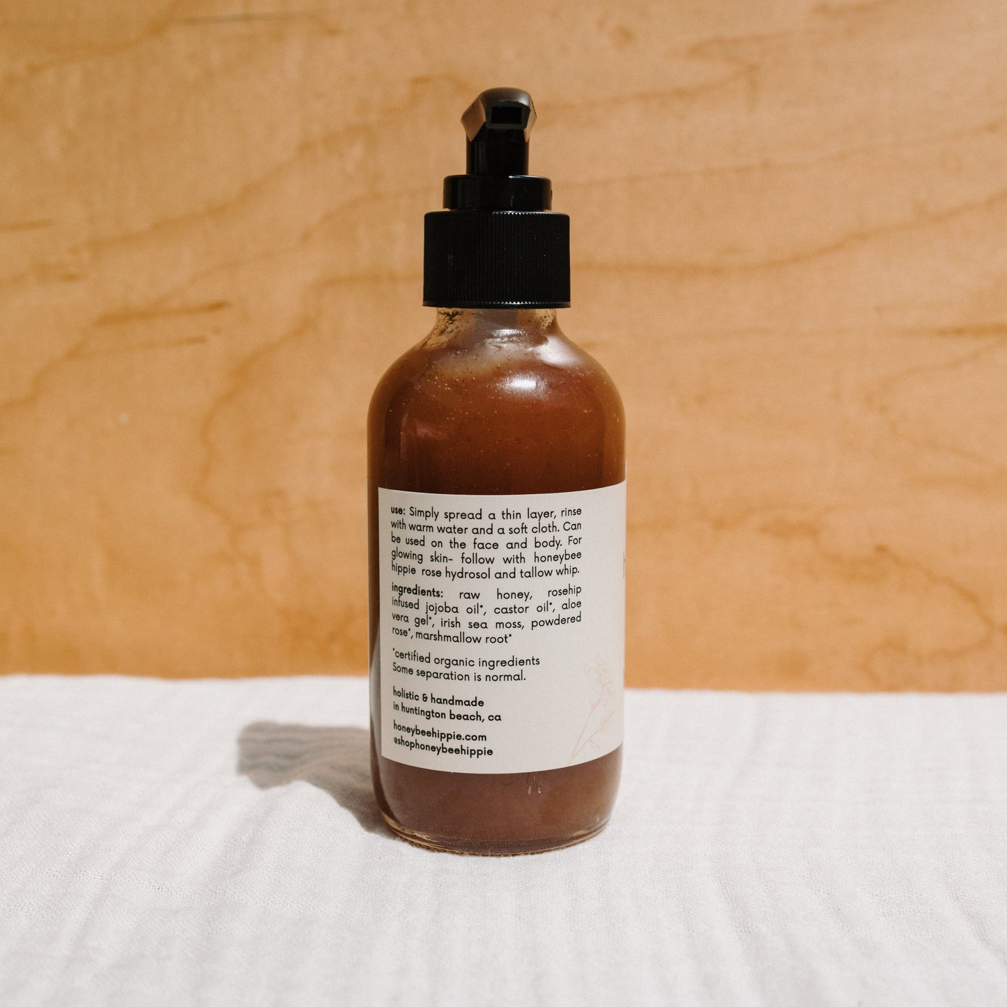 A brown glass pump bottle labeled with "Honey Glow Herbal Honey & Oil Wash" from Honeybee Hippie sits against a wooden background, highlighting its organic skincare origins and clear application instructions for optimal results.