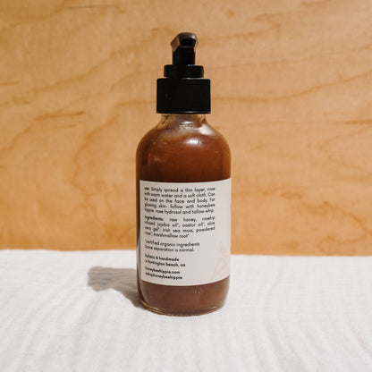A brown glass pump bottle labeled with "Honey Glow Herbal Honey & Oil Wash" from Honeybee Hippie sits against a wooden background, highlighting its organic skincare origins and clear application instructions for optimal results.