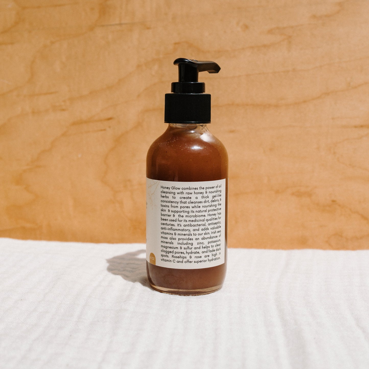 The Honeybee Hippie "Honey Glow" Herbal Honey & Oil Wash, an organic skincare product with a pump dispenser, sits on a textured surface. Against a wooden backdrop, its brown honey-infused liquid is highlighted by the text-filled label promoting its natural facial cleansing benefits.