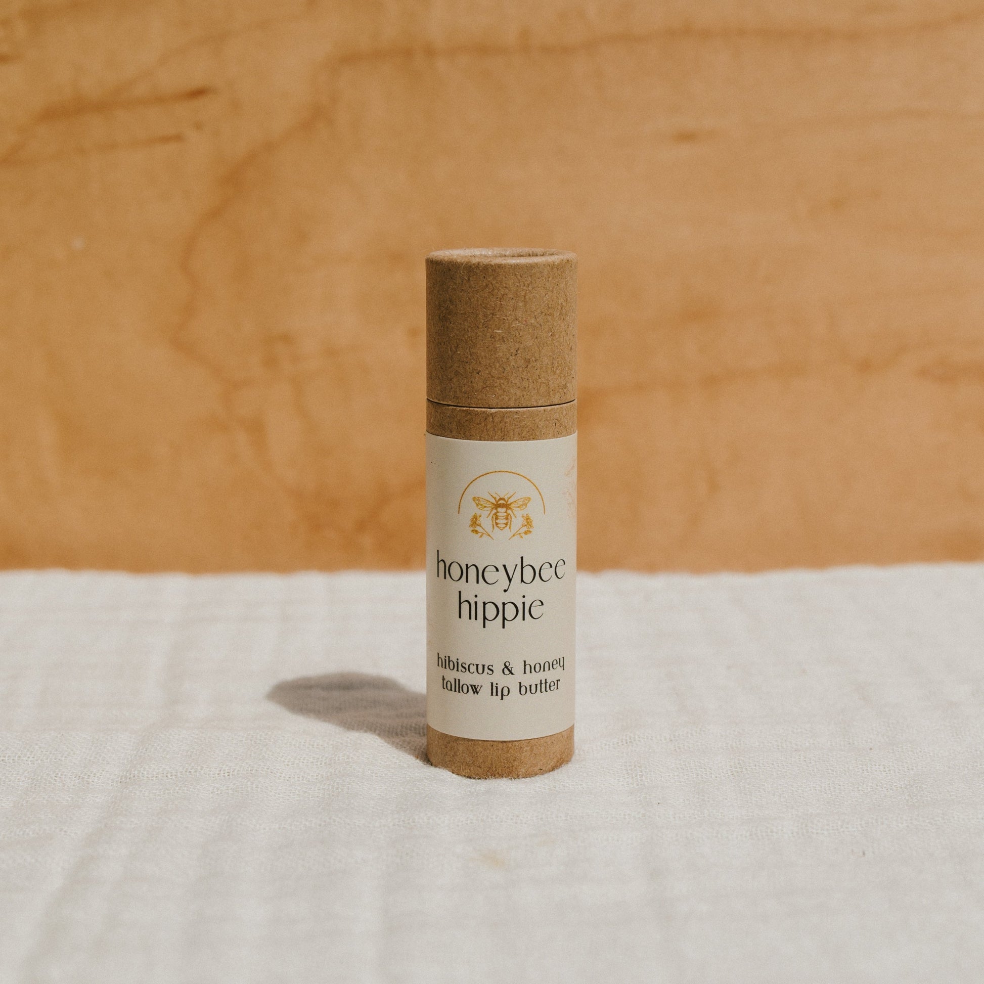 A cylindrical cardboard tube labeled "Honeybee Hippie" stands upright, displaying a bee illustration and described as "hibiscus & honey tallow lip butter." It rests on textured, light fabric against a wooden background.