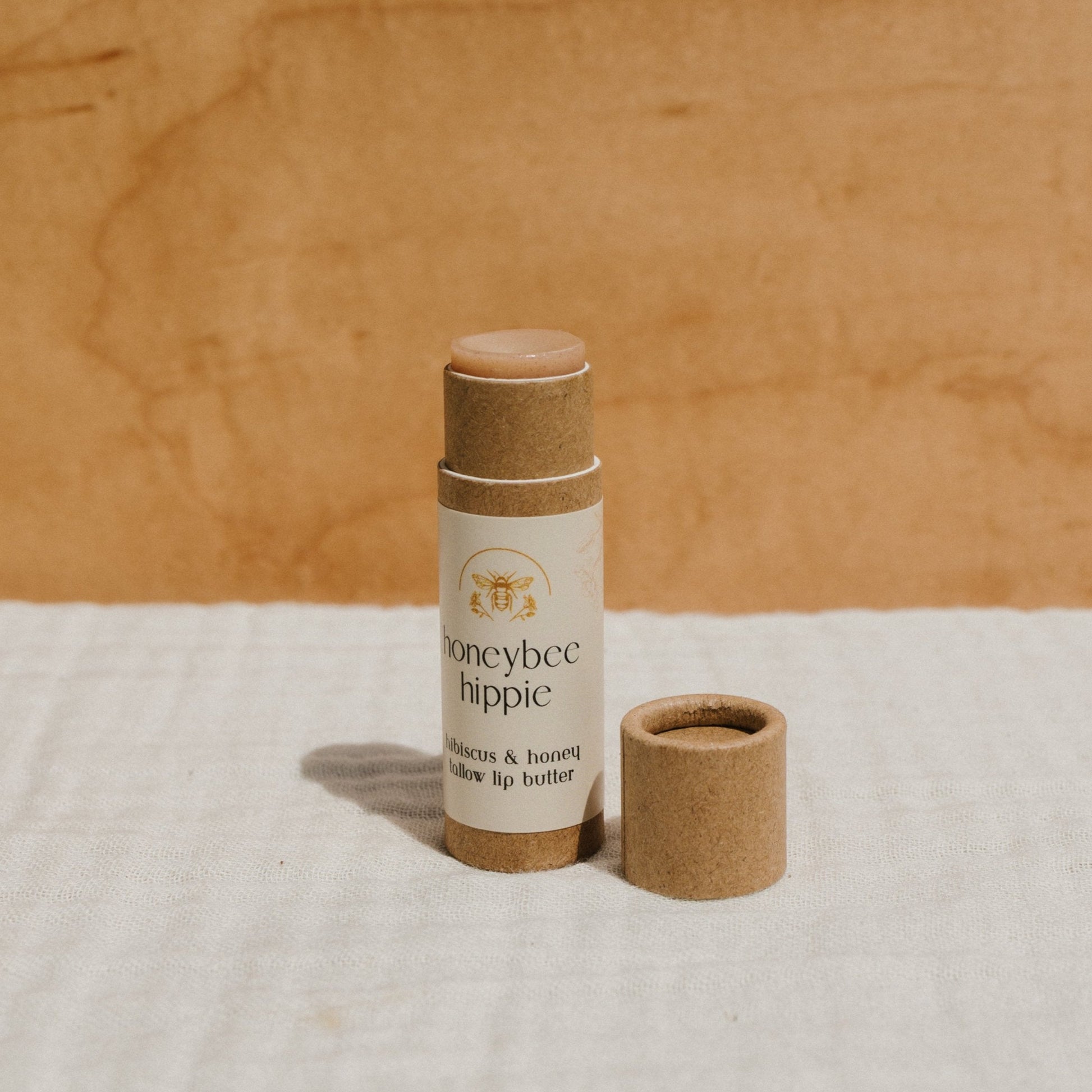 A tube of Honeybee Hippie's hibiscus & honey tallow lip butter stands upright with its cap beside it on a white textured surface against a warm beige background.