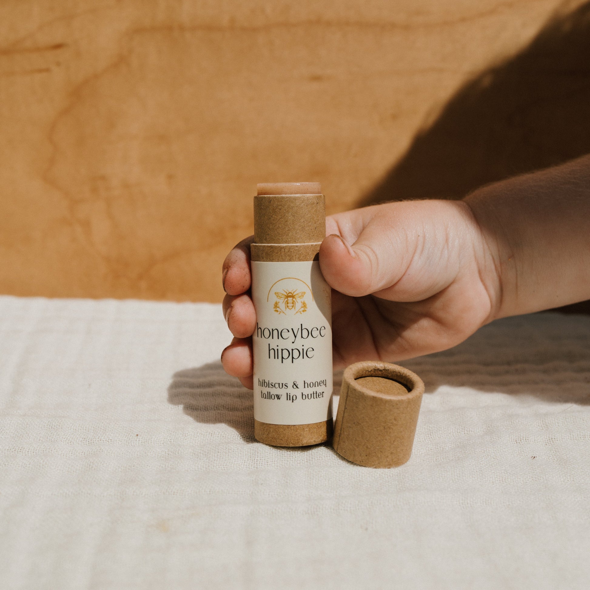 A hand holds a Honeybee Hippie hibiscus & honey tallow lip butter, enriched with organic herbs. The eco-friendly tube has a beige cap resting beside it, set against a light fabric backdrop and wooden surface.