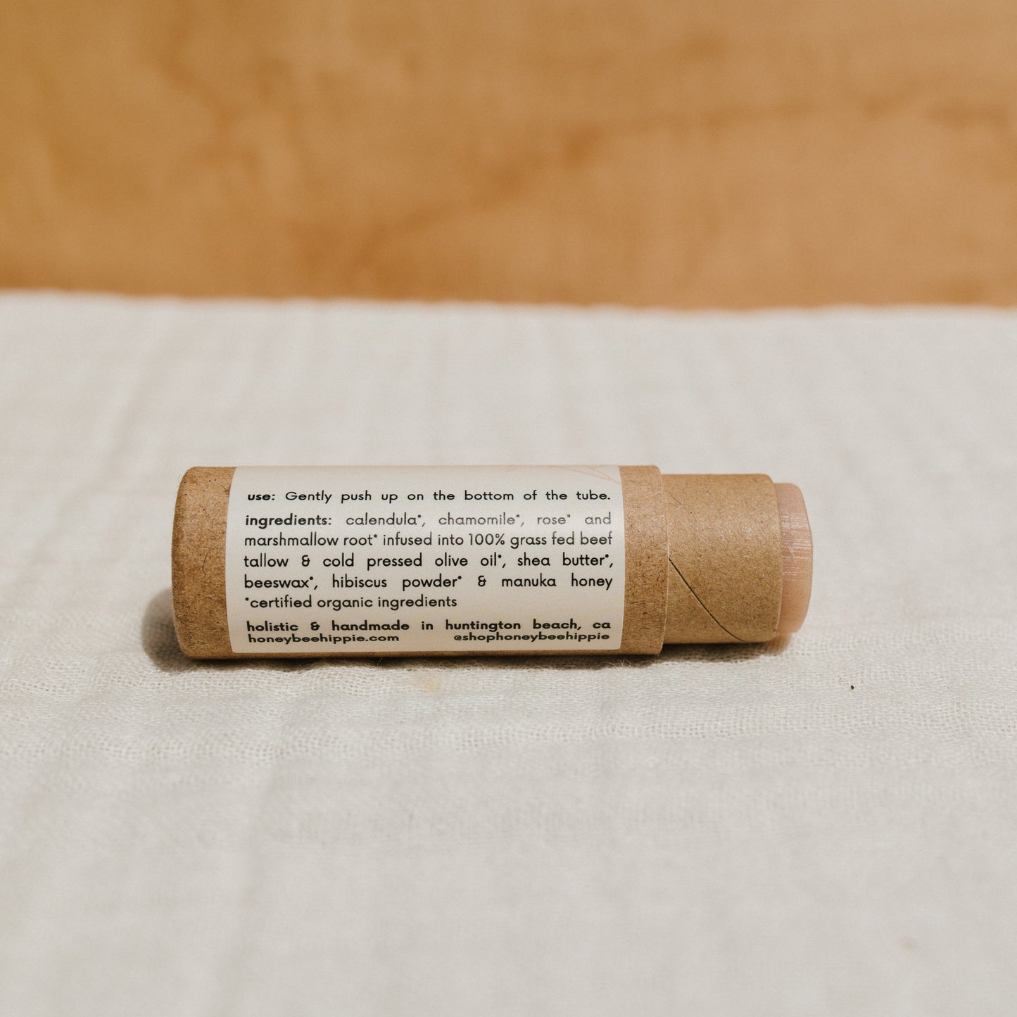 Close-up of Honeybee Hippie's hibiscus & honey tallow lip butter in a cardboard tube against a beige backdrop. The label highlights moisturizing ingredients like calendula, chamomile, shea butter, and hibiscus powder, with instructions to push up from the bottom.
