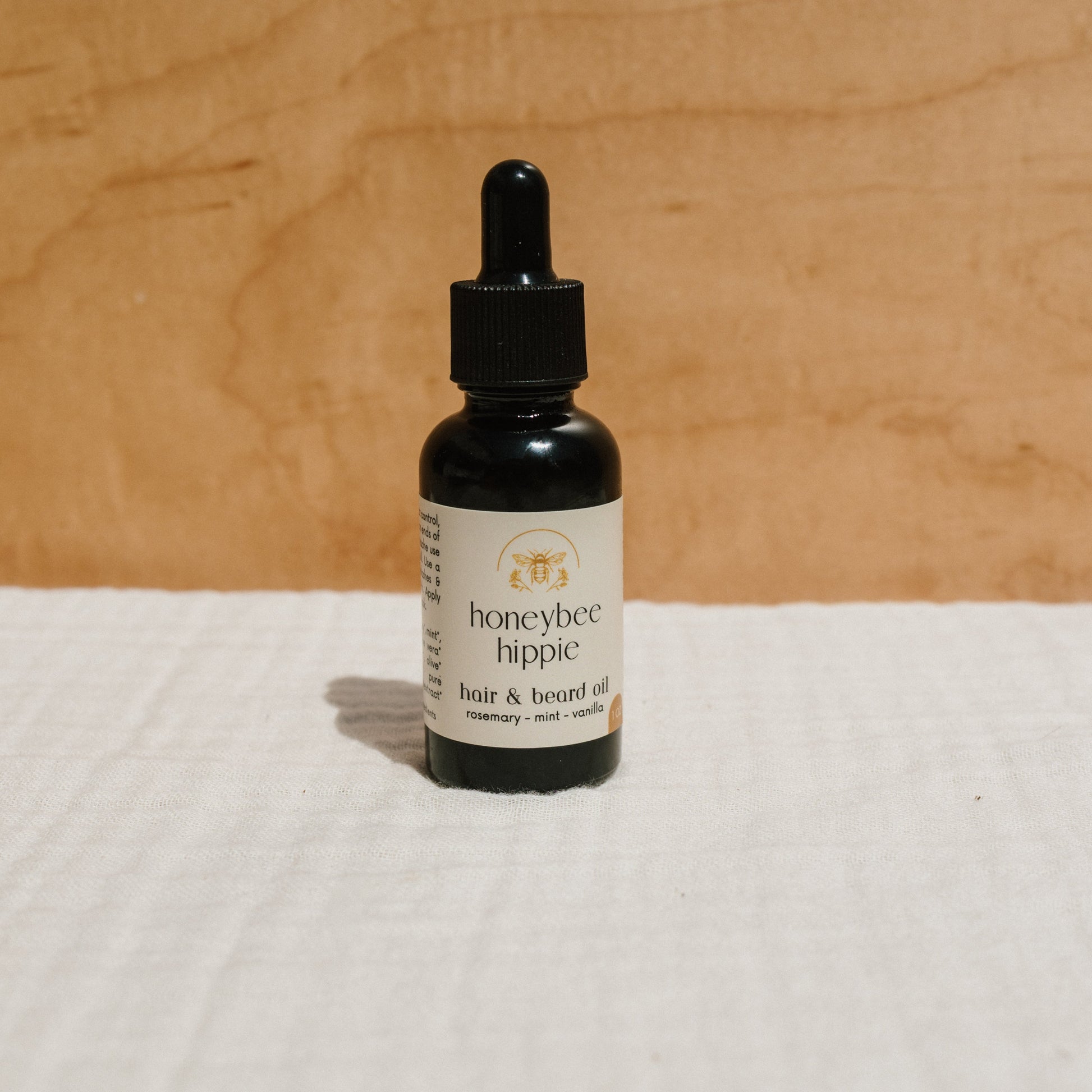 A dark dropper bottle labeled "Honeybee Hippie" contains organic hair & beard oil with rosemary, mint, and vanilla for frizz control. It's elegantly placed on white fabric against a wooden background.
