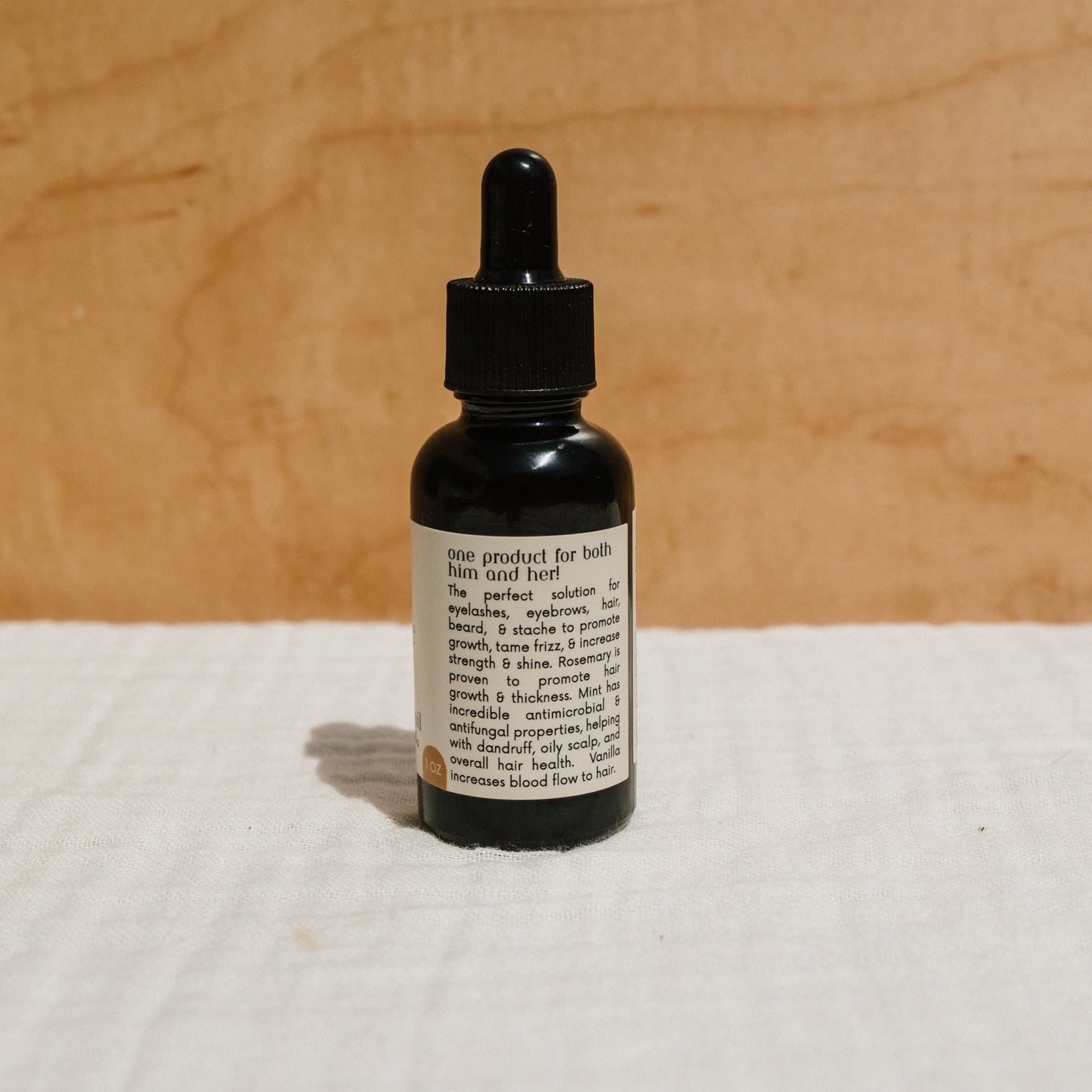 A small bottle with a black dropper cap rests on a textured white surface. The label shows it's Honeybee Hippie's hair & beard oil, an organic product for thin and thick hair that promotes growth and boosts scalp blood flow. Perfect for frizz control, it blends into any neutral setting effortlessly.