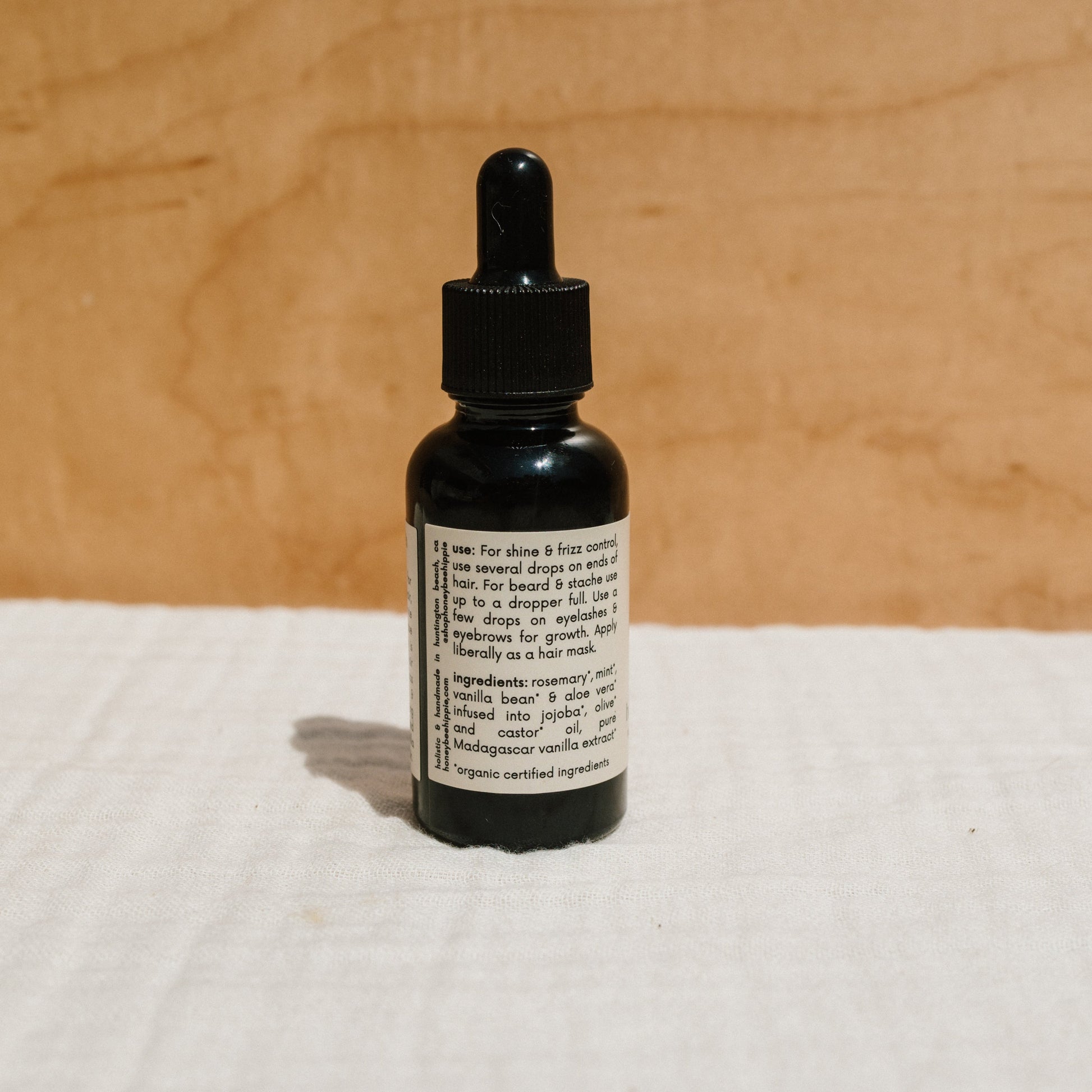 A small brown bottle of Honeybee Hippie's hair & beard oil with a black dropper cap rests on a beige surface. The label, highlighting frizz control benefits and ingredients, blends seamlessly with the warm wooden background.