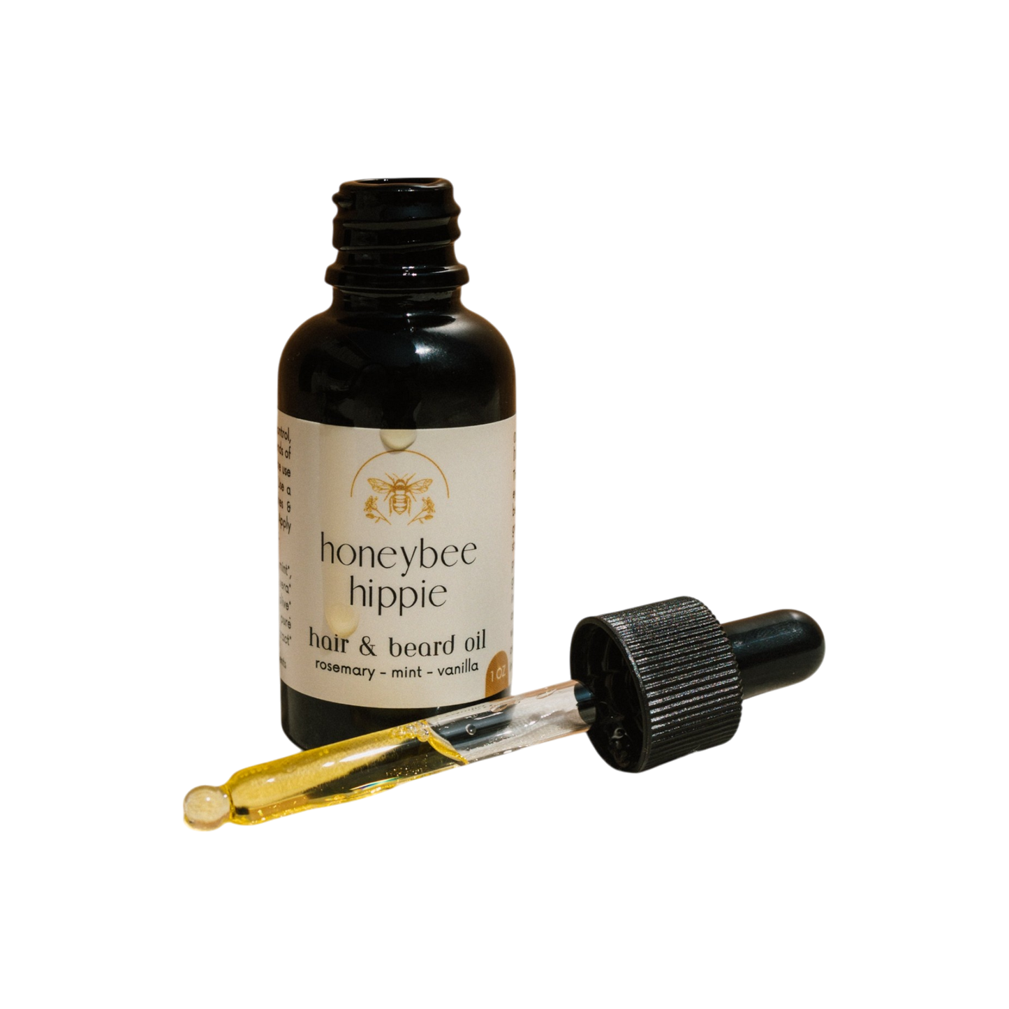 A black Honeybee Hippie hair & beard oil dropper bottle is shown, featuring organic oils. The golden oil dropper is placed in front, highlighting rosemary, mint, and vanilla on the label—ideal for healthy hair growth.