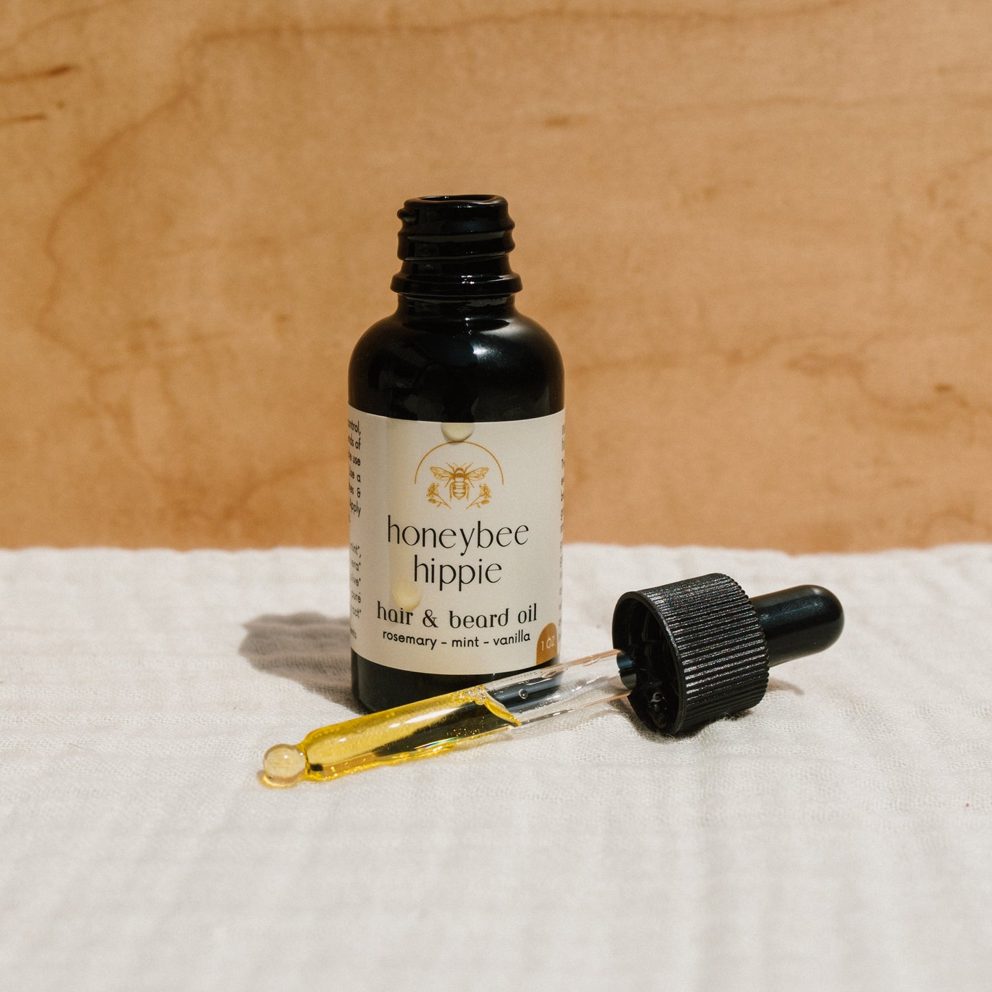 A Honeybee Hippie hair & beard oil bottle with a dropper rests on a white textured cloth. The rosemary, mint, and vanilla label promises nourishment and growth. Its warm brown packaging enhances its pledge for better hair health.