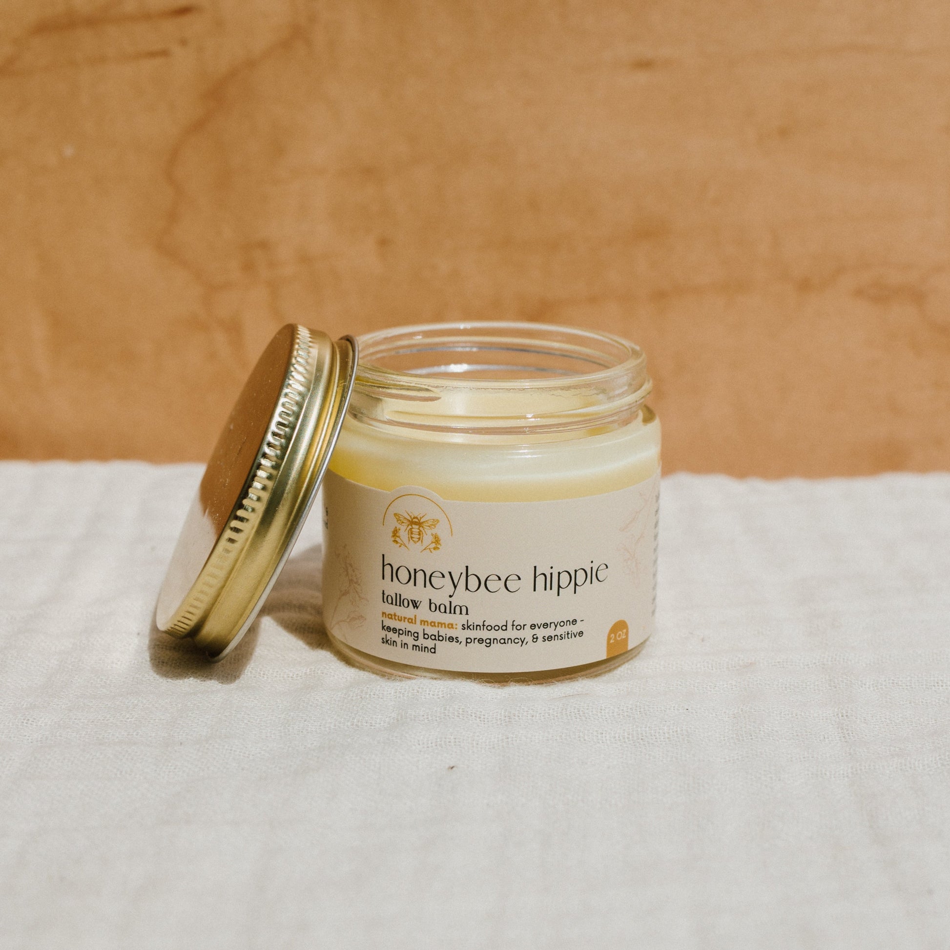 A jar of Honeybee Hippie's natural mama tallow balm, with a metal lid partially open, rests on light fabric against a wooden backdrop. Its label confirms safety for babies, pregnancy, and sensitive skin. Made from grass-fed tallow, it effectively soothes skin irritations.
