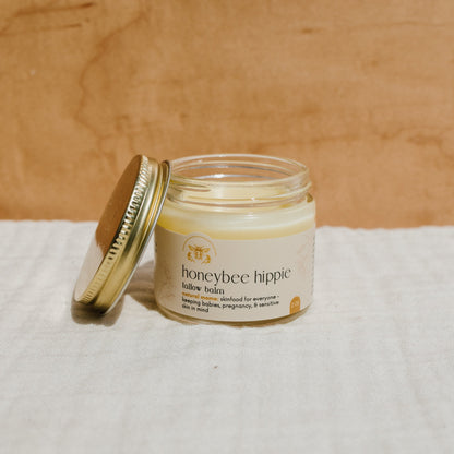 A jar of Honeybee Hippie's natural mama tallow balm, with a metal lid partially open, rests on light fabric against a wooden backdrop. Its label confirms safety for babies, pregnancy, and sensitive skin. Made from grass-fed tallow, it effectively soothes skin irritations.