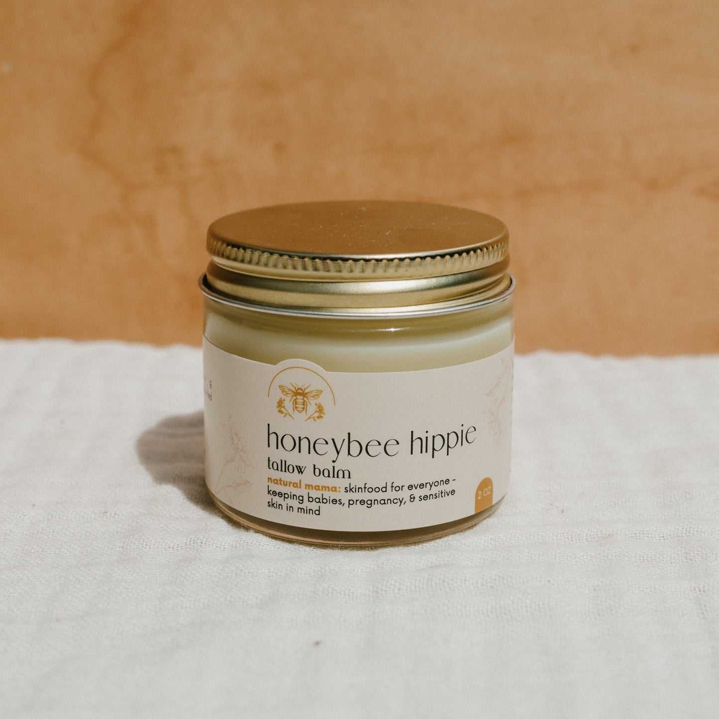 A jar of Honeybee Hippie's natural mama tallow balm sits on a white cloth against a beige background, with a label featuring grass-fed tallow and organic herbs. This natural skincare product is ideal for everyone, including babies and pregnant women, providing eczema relief.