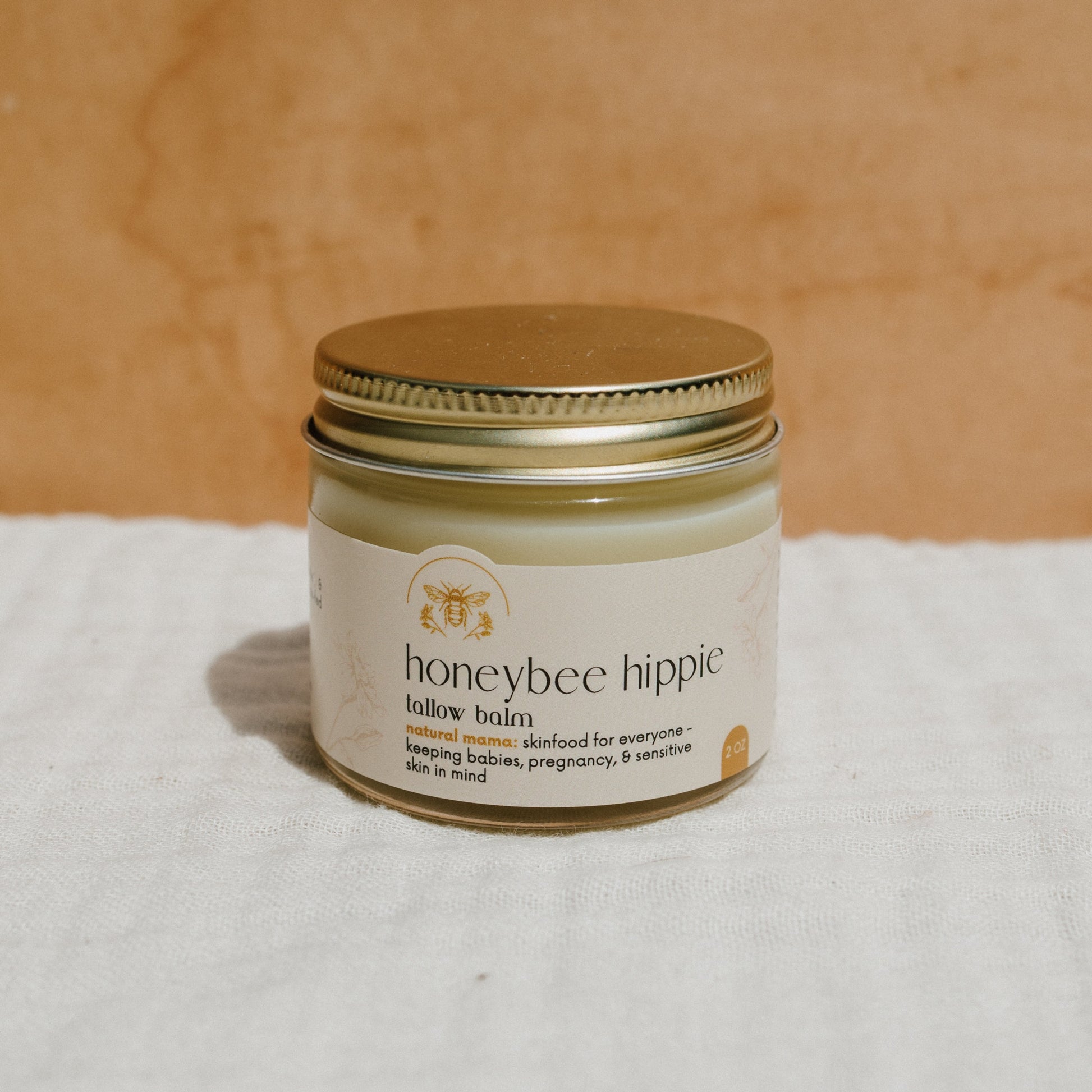 A jar of Honeybee Hippie's natural mama tallow balm sits on a white cloth against a beige background, with a label featuring grass-fed tallow and organic herbs. This natural skincare product is ideal for everyone, including babies and pregnant women, providing eczema relief.