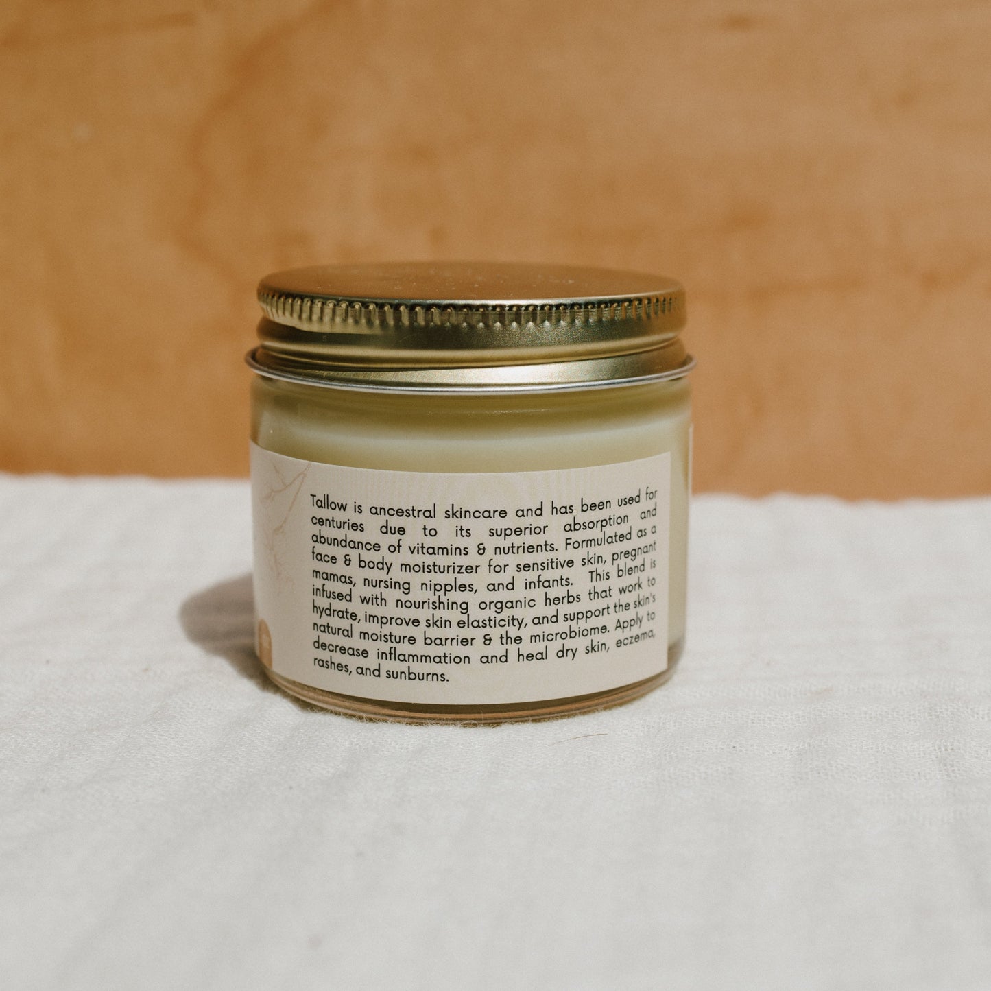 A small jar with a metal lid sits on a white cloth, labeled as "natural mama tallow balm" by Honeybee Hippie. It claims to soothe skin irritations, dry patches, rashes, and sunburns. The background is a warm brown surface.