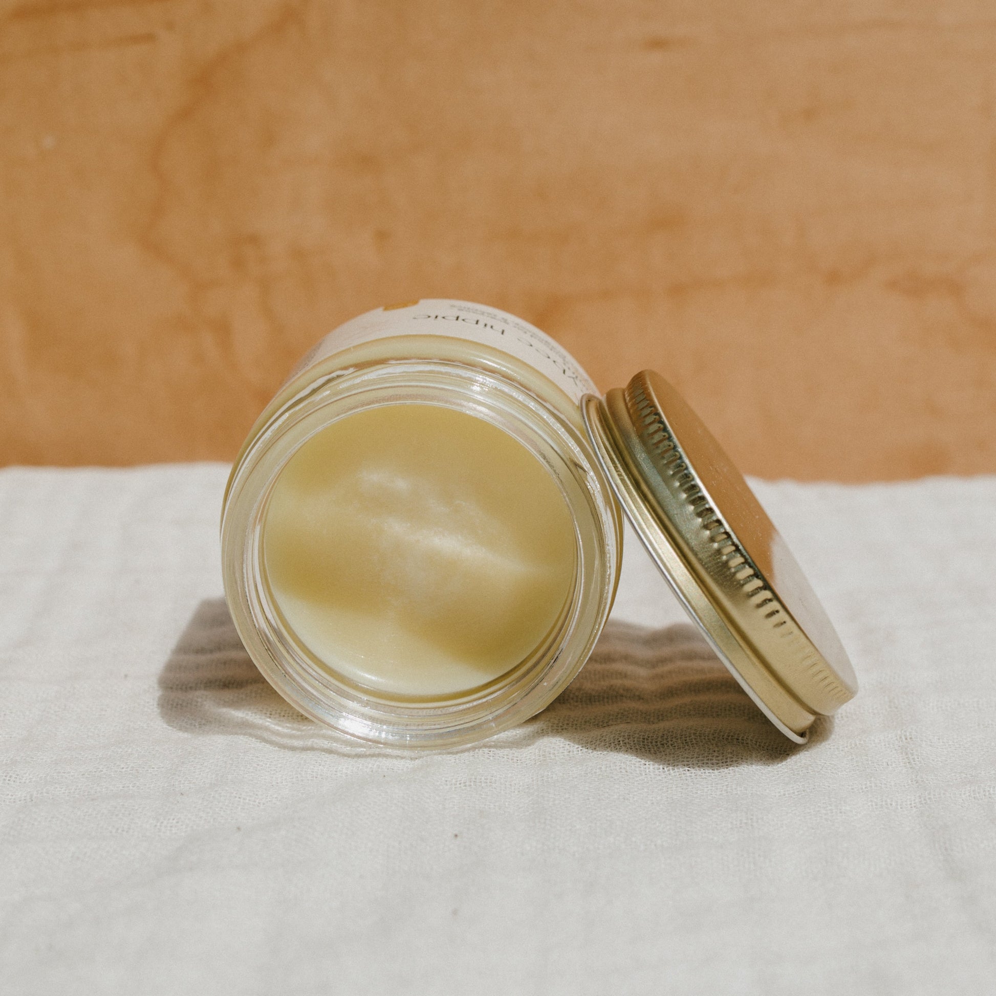 An open glass jar with a metal lid sits on a white cloth, showcasing Honeybee Hippie's natural mama tallow balm—a creamy, wax-like skin moisturizer made from grass-fed tallow—against a light brown wood surface.