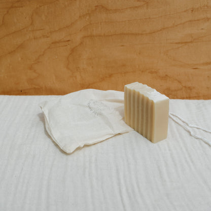 A Honeybee Hippie tallow soap bar stands upright on white fabric. Nearby, a crumpled drawstring bag filled with organic herbs lies beside it against a wooden panel backdrop, creating a simple, natural setting.