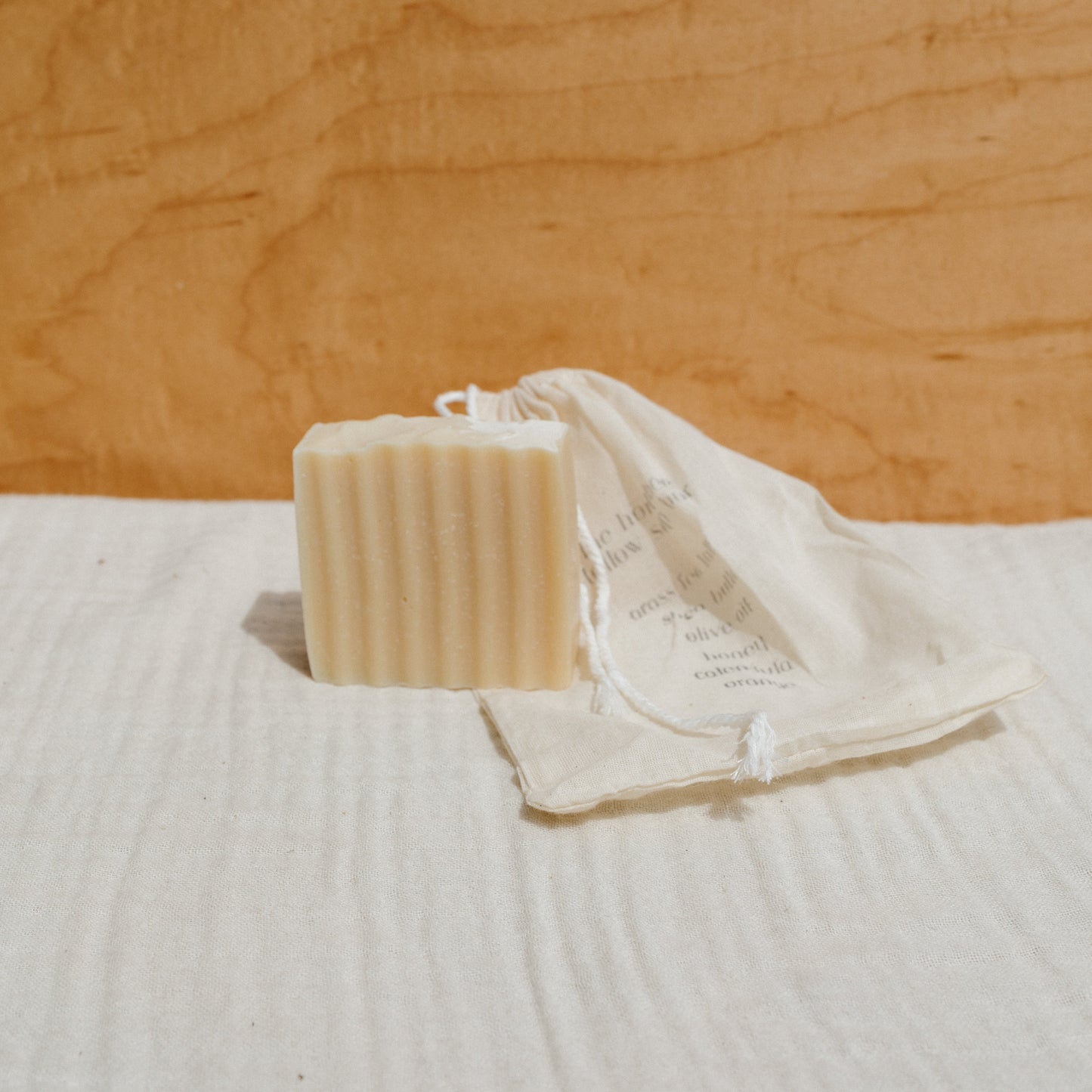 A ridged bar of Honeybee Hippie's the honeybee tallow soap rests on a white cloth next to a partially open, printed cotton drawstring bag, set against a light wood background for a clean, natural look infused with organic herbs.