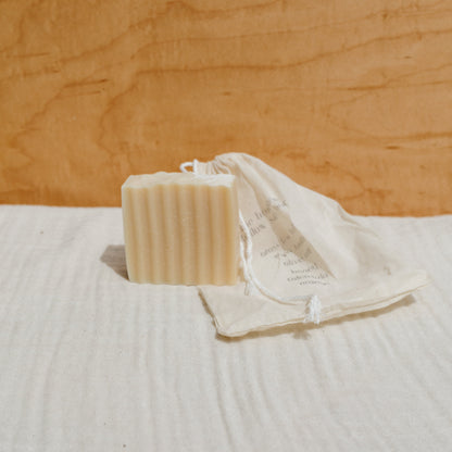 A ridged bar of Honeybee Hippie's the honeybee tallow soap rests on a white cloth next to a partially open, printed cotton drawstring bag, set against a light wood background for a clean, natural look infused with organic herbs.