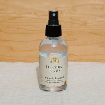 A bottle of Honeybee Hippie's butterfly hydrosol with spray top, set against a wooden background, features steam-distilled organic helichrysum and butterfly pea flower for a serene blend.