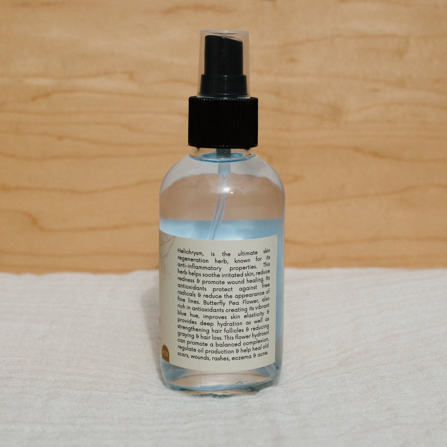 A clear Butterfly Hydrosol by Honeybee Hippie, with a black cap and detailed label, sits on a soft, white surface against a warm wooden background.