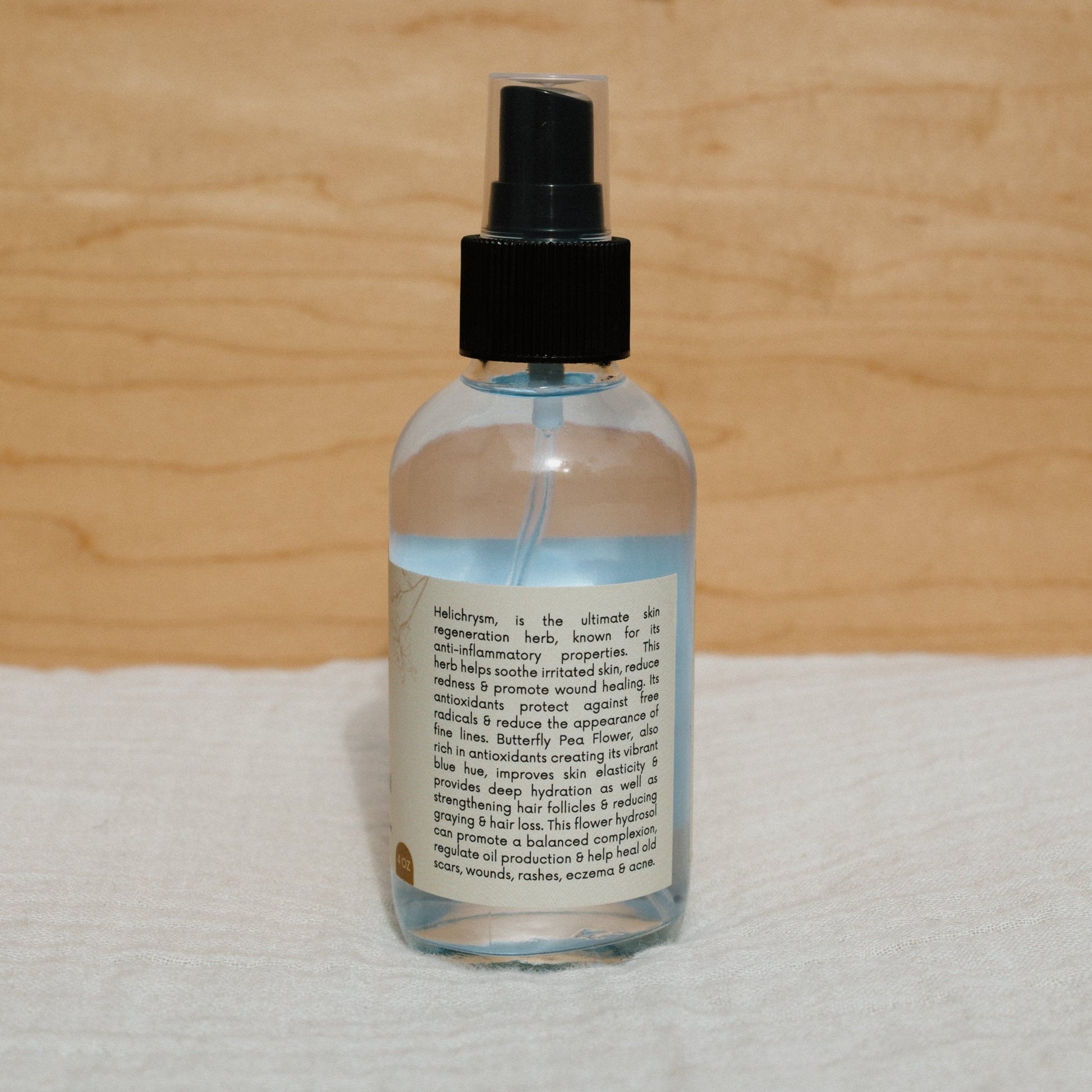 A clear Butterfly Hydrosol by Honeybee Hippie, with a black cap and detailed label, sits on a soft, white surface against a warm wooden background.