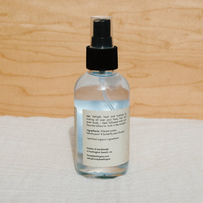 A Butterfly Hydrosol spray bottle from Honeybee Hippie features a black nozzle and label with usage instructions and ingredients like filtered water and helichrysm. The bottle rests elegantly on a soft, light-colored surface.