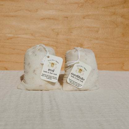 Two small drawstring bags, labeled "rest" and "meditate", rest on a light fabric surface. The meditate bath soak by Honeybee Hippie contains soothing bath salts, with a tag featuring an organic flower design, set against a wooden backdrop.
