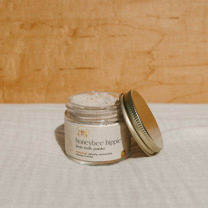 A Honeybee Hippie "fresh tooth powder" jar with an open lid sits on a white cloth, partially filled with white powder. It features a simple label promising to rebuild tooth enamel against a backdrop of light wooden texture.