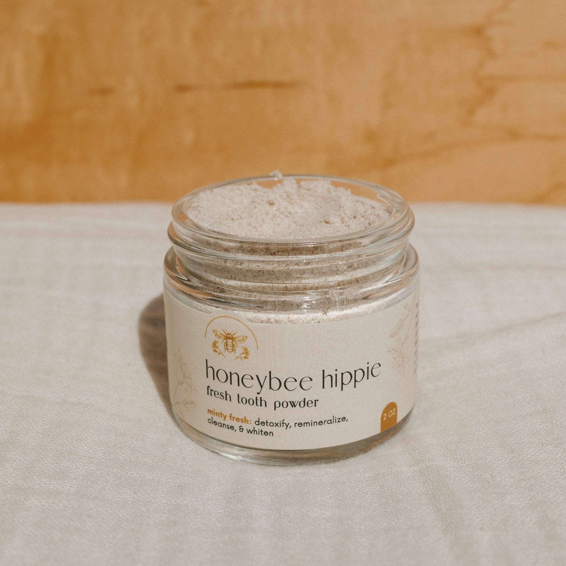 A glass jar of Honeybee Hippie's "fresh tooth powder" rests on a light fabric. The label highlights its features: minty fresh, detoxify, remineralize, cleanse, and whiten. A warm, textured wooden background completes the scene.