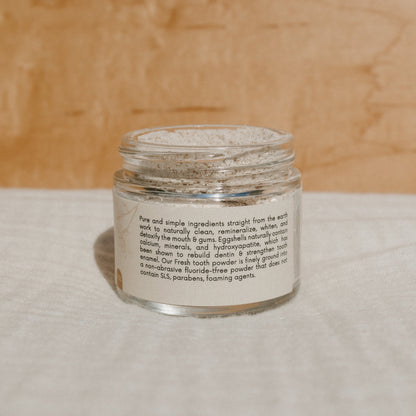A clear glass jar with a beige lid contains Honeybee Hippie's fresh tooth powder, featuring natural ingredients and a fluoride-free formula that remineralizes teeth. Resting on soft, light-colored fabric, it exudes purity and wellness.