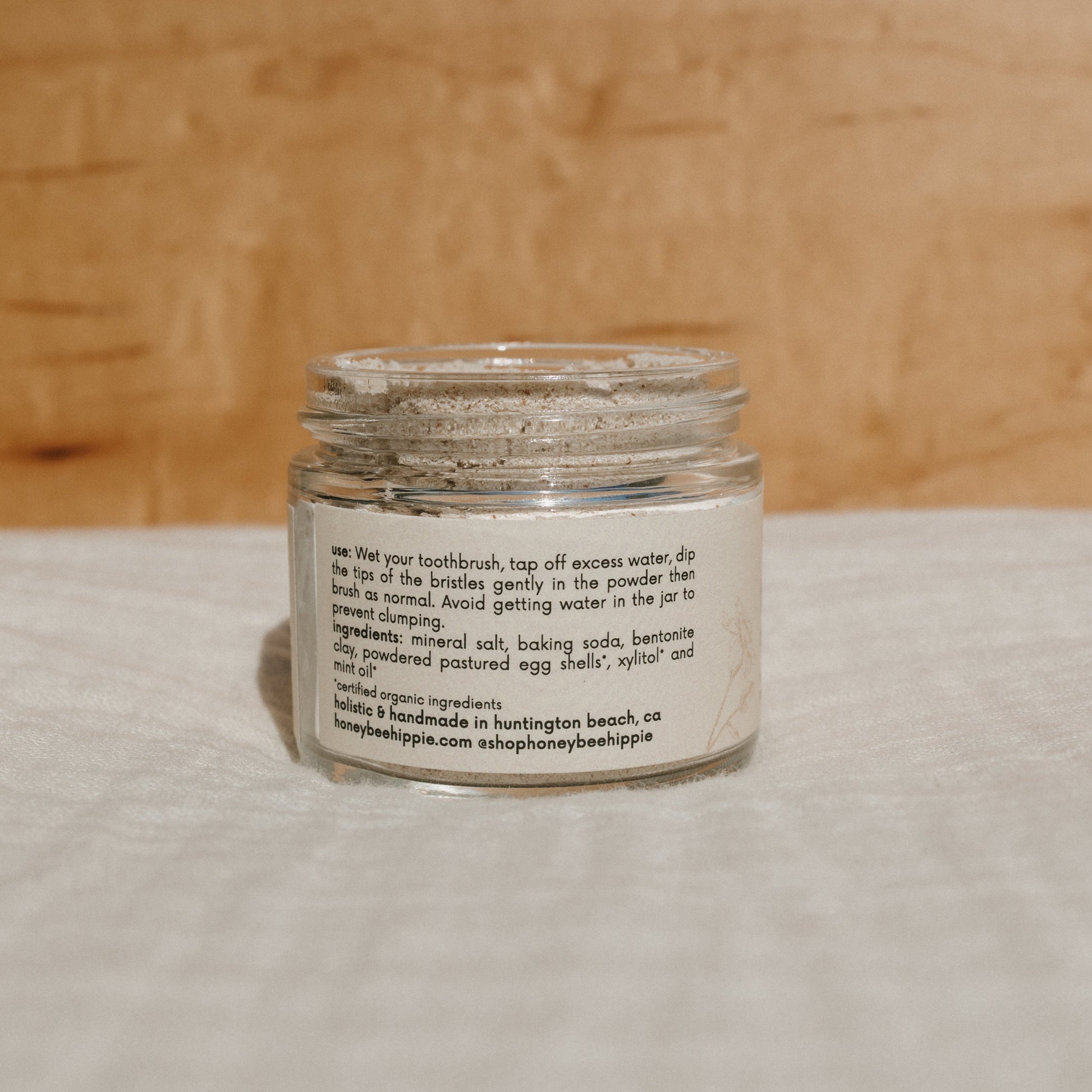 A glass jar of fresh tooth powder from Honeybee Hippie rests on a wooden surface. The label features usage instructions and lists mineral salt and baking soda as key ingredients to remineralize teeth, along with Honeybee Hippie's contact information.