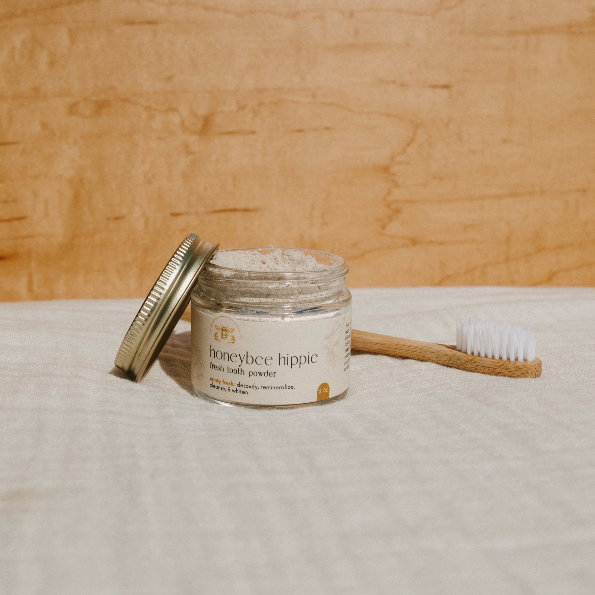 A "Honeybee Hippie fresh tooth powder" jar, known for remineralizing teeth, sits with its lid against it. Nearby, a wooden toothbrush with white bristles rests on pale fabric against a light wood background.