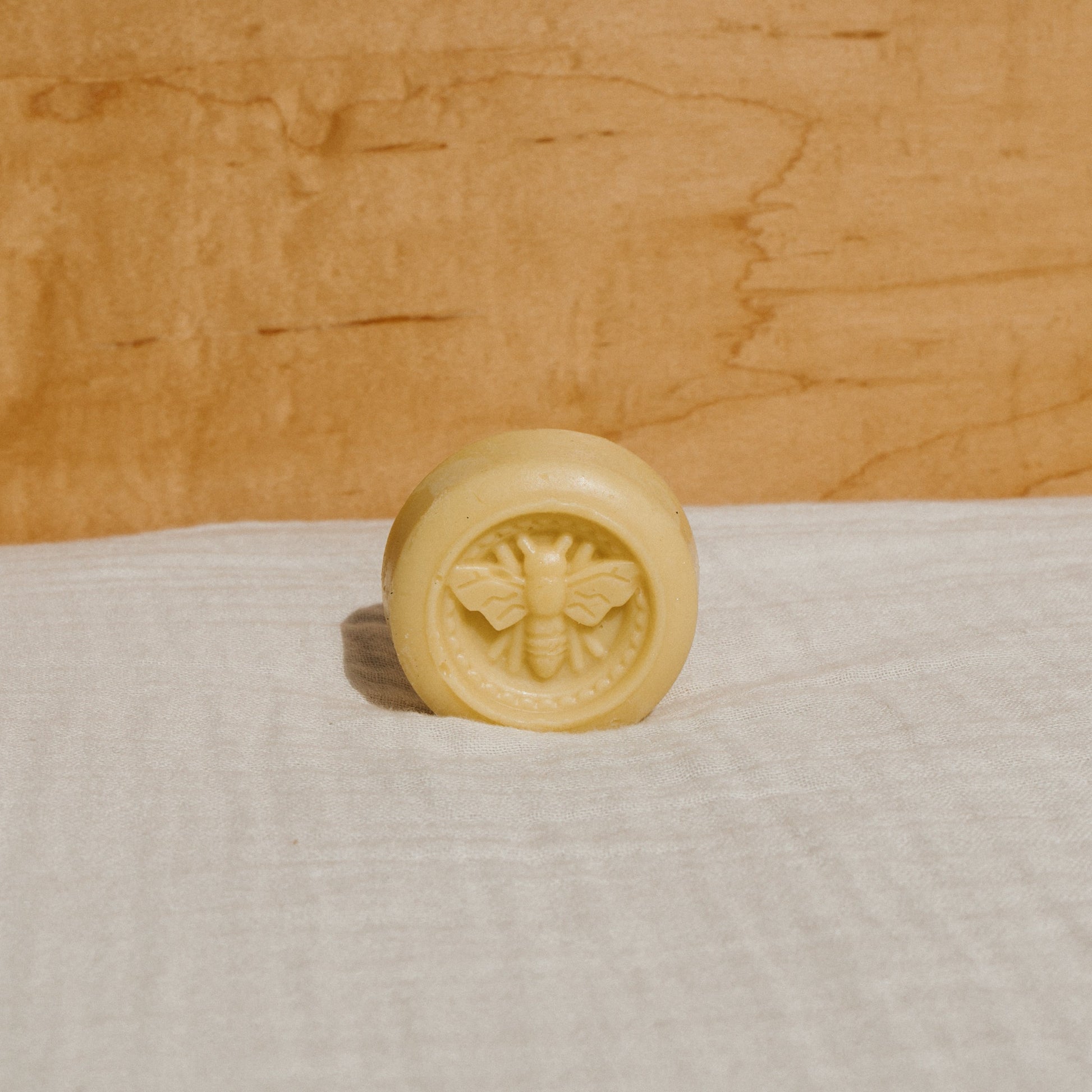 A round, yellow tallow lotion bar by Honeybee Hippie, embossed with a bee design, is placed on soft white fabric. Its natural moisturizer qualities are accented by the warm, light brown wood background.