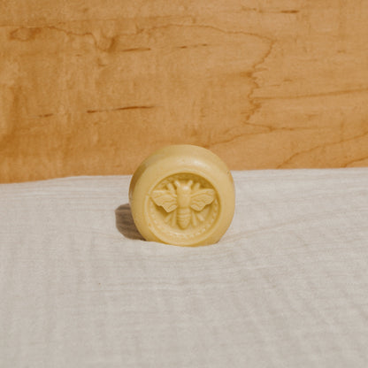 A round, yellow tallow lotion bar by Honeybee Hippie, embossed with a bee design, is placed on soft white fabric. Its natural moisturizer qualities are accented by the warm, light brown wood background.