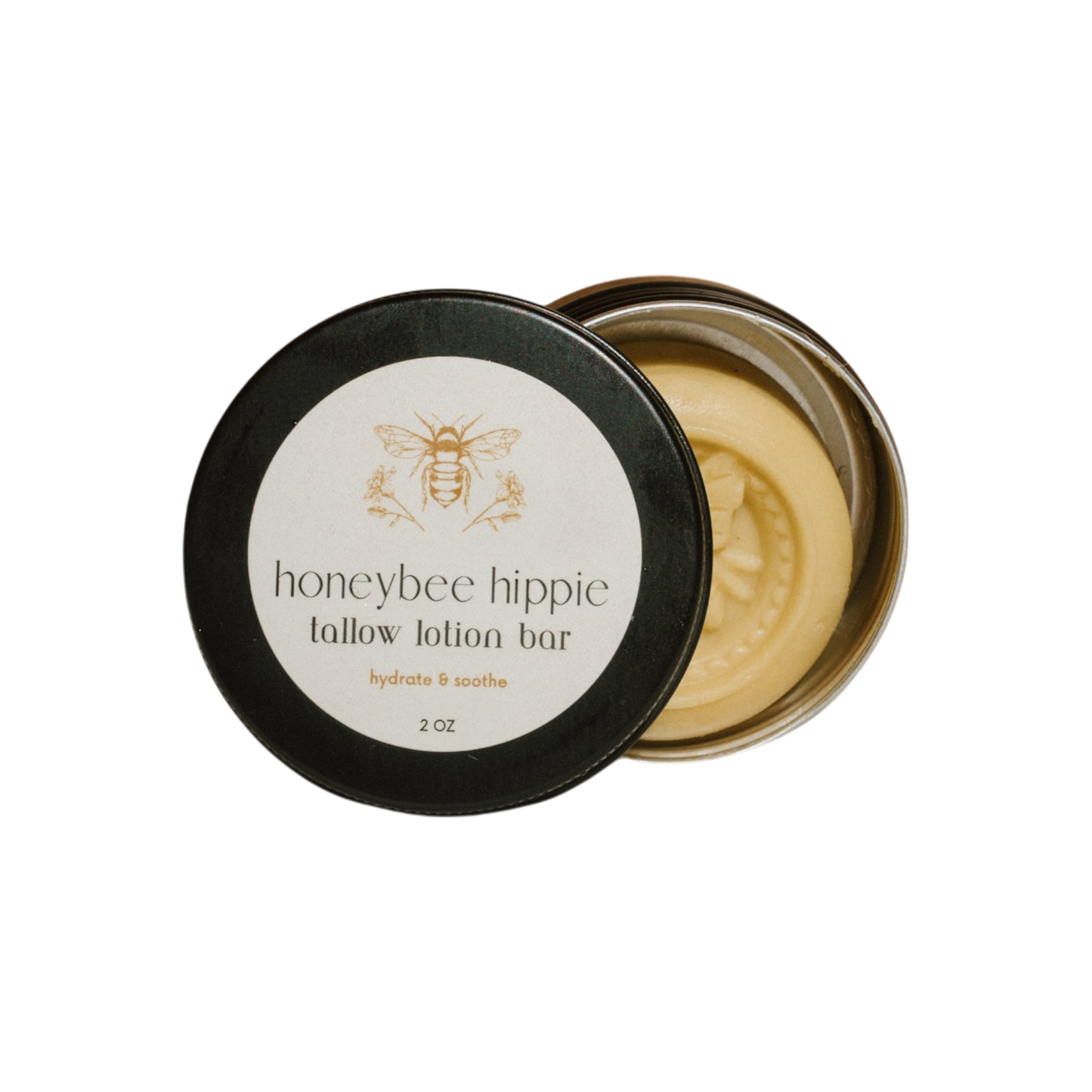 A round tin with an open lid reveals a Honeybee Hippie tallow lotion bar made from grass-fed tallow, featuring a bee logo and "tallow lotion bar, hydrate & soothe, 2 oz." on the lid. The moisturizer is adorned with a decorative bee imprint.