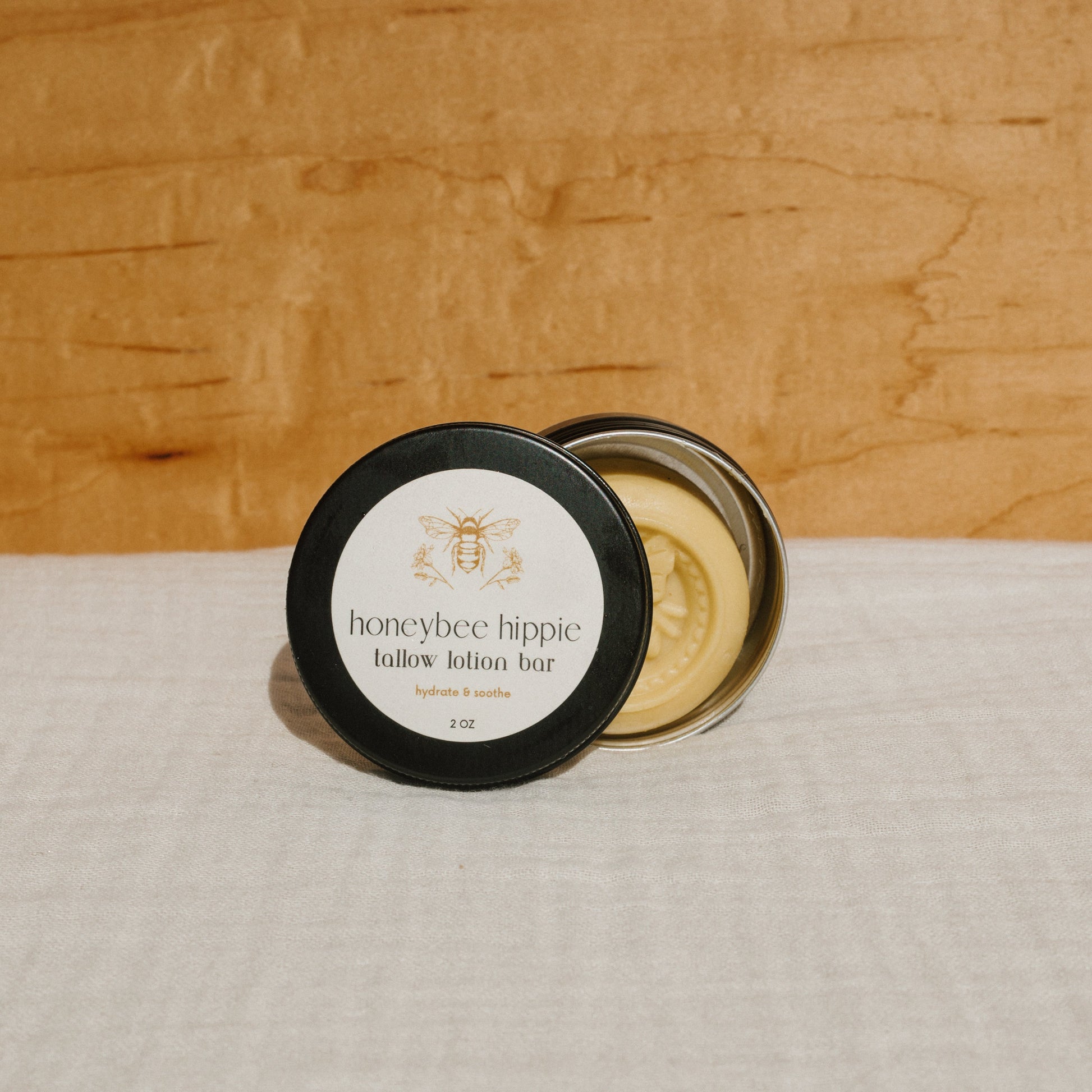 A round tin labeled "Honeybee Hippie tallow lotion bar" with a black lid, shown open to display the lotion bar infused with organic herbs, rests on a light-colored fabric surface against a wooden background.
