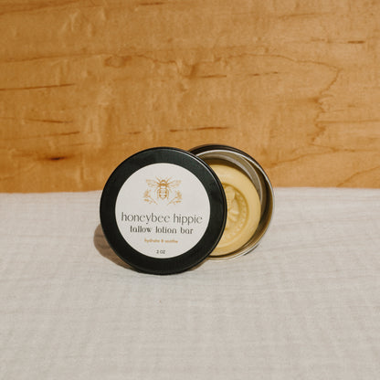 A round tin labeled "Honeybee Hippie tallow lotion bar" with a black lid, shown open to display the lotion bar infused with organic herbs, rests on a light-colored fabric surface against a wooden background.