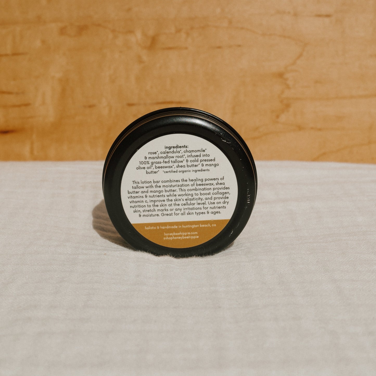 A small round tallow lotion bar by Honeybee Hippie, enriched with rose, chamomile, and shea butter, sits on a wooden background. The label highlights its moisturizing benefits for all skin types and ages using the nourishing power of grass-fed tallow.