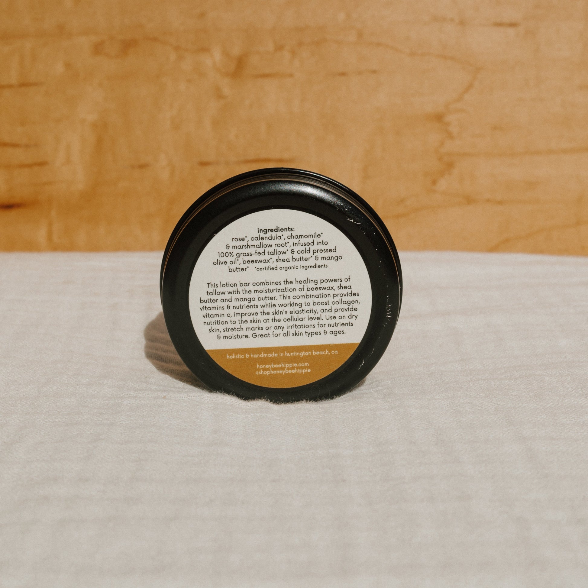 A small round tallow lotion bar by Honeybee Hippie, enriched with rose, chamomile, and shea butter, sits on a wooden background. The label highlights its moisturizing benefits for all skin types and ages using the nourishing power of grass-fed tallow.