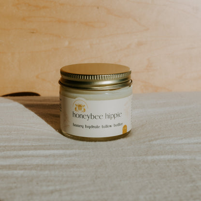 A jar of Honeybee Hippie's honey hydrate tallow butter rests on a light surface enriched with organic herbs. The label displays a bee illustration, and the metallic gold lid shines against the smooth wooden backdrop.